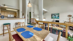 Une cuisine moderne équipée d'un four à micro-ondes, d'un réfrigérateur et d'un coin bar. On y trouve une table à manger avec des sets de table bleus et des chaises en bois. Un escalier mène à un espace de vie comprenant un canapé et un tableau accroché au mur.