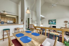 Eine moderne Küche mit Mikrowelle, Kühlschrank und einer Bar. Ein Esstisch mit blauen Unterlagen und Holzkästen. Eine Treppe führt zu einem Wohnbereich mit einer Couch und einem Bild an der Wand.
