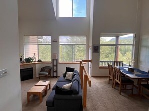 Ein geräumiges Wohnzimmer mit einer Couch, einem Couchtisch und einem Essbereich mit Stühlen und einem Tisch. Große Fenster bieten Tageslicht und Blick ins Freie.