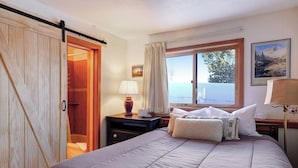 Quarto com cama, mesa de cabeceira, abajur, janela com cortinas e porta para outro cômodo.