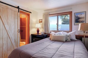 Ein Schlafzimmer mit einem Bett, einem Nachttisch, einer Lampe, einem Fenster mit Vorhängen und einer Tür, die in einen anderen Raum führt.