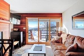 Ein gemütliches Wohnzimmer mit einer Couch, einem Couchtisch und einem Fernseher. Eine Glasschiebetür führt in einen verschneiten Außenbereich.