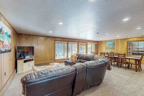A living room with a sectional sofa, a television, a dining table with chairs, and large windows. - Spacious cabin with river views, private hot tub, sun room, and full kitchen! (South Fork)