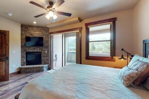Ein Schlafzimmer mit Kamin aus Stein, Deckenventilator, einem großen Bett und einer Glastür, die auf einen Balkon führt.