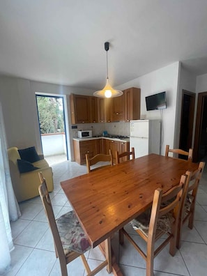 A dining area with a wooden table and chairs, a kitchen with wooden cabinets, a microwave, and a TV mounted on the wall. - Holiday apartment with air conditioning and balcony (Rosolina Mare)