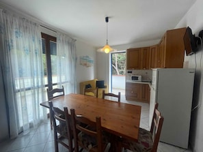 A dining area with a wooden table and chairs, a kitchen with wooden cabinets and appliances, and a window with curtains. - Holiday apartment with air conditioning and balcony (Rosolina Mare)
