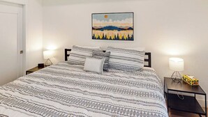 Una cama bien hecha con ropa de cama estampada, un cuadro colgado en la pared y lámparas de noche sobre una mesita.
