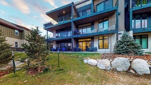 Un edificio de varios pisos con balcones, un pequeño jardín con rocas y un árbol, y un cielo despejado.