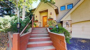 Une maison d’habitation avec un escalier en bois menant à la porte d’entrée, entourée de verdure et d’un mur en pierre.