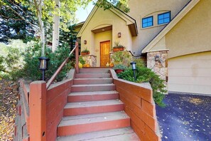 Ein Wohnhaus mit einer Holztreppe, die zu einer Haustür führt, umgeben von Grünpflanzen und einer Steinmauer.