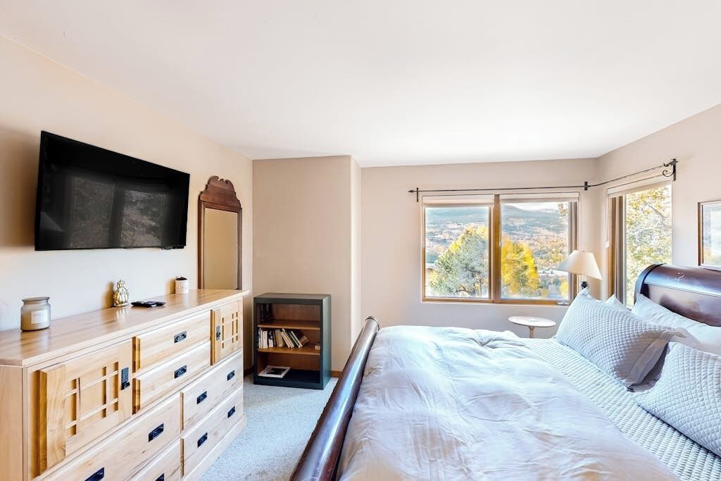 A bedroom with a bed, dresser, TV, bookshelf, and large windows with a view.