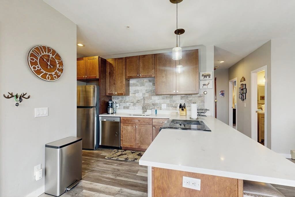 A modern kitchen with wooden cabinets, a stainless steel refrigerator, a large island, and a wall clock.
