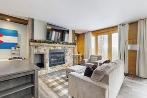 A modern living room with a stone fireplace, a sofa, a coffee table, and a TV mounted on the wall. - Cozy condo with fireplace, pool, & hot tub - near trails (Silverthorne)