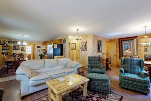 Ein gemütliches Wohnzimmer mit einer Couch, zwei karierten Sesseln, einem Couchtisch, Holzwänden, einem Essbereich mit Tisch und Stühlen sowie einer Küchenzeile mit Kühlschrank.