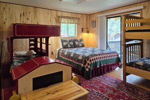 Ein Zimmer mit Etagenbetten, einem Bett mit Quilt, einem Holztisch, einem Stuhl, einem Fenster mit Vorhängen und einer Tür, die nach draußen führt.