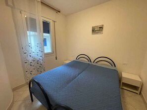 A bedroom with a bed, a window with curtains, a small bedside table, and a wall-mounted picture. - Holiday apartment with air conditioning and balcony (Rosolina Mare)