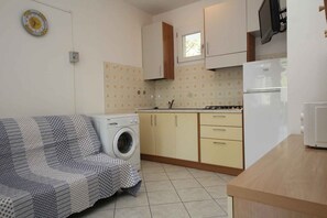 A compact kitchen with a washing machine, a dining area with a patterned sofa, and a small window. - Holiday apartment with air conditioning and balcony (Rosolina Mare)