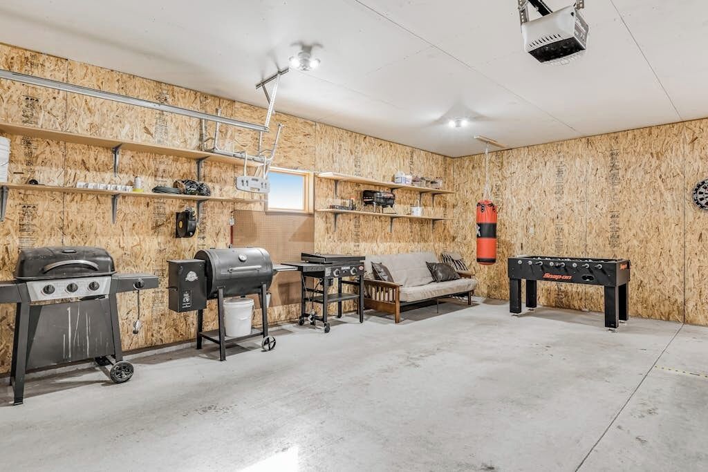 A room with a concrete floor, wooden walls, a sofa, a table, a grill, a projector, and shelves with various items.