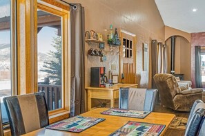 Ein gemütliches Interieur mit einem Holzespeisetisch, Ledersesseln, einer Kaffeemaschine und Blick auf verschneite Berge durch große Fenster.