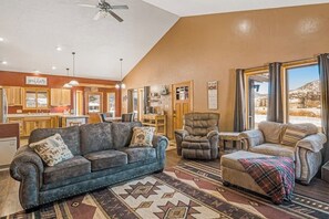 Ein großzügiger Wohnbereich mit Sofa, Sessel, Hocker und Teppich. Im Hintergrund befindet sich eine Küche mit Holzschränken und einem Essbereich.