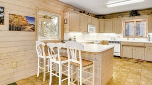 Une cuisine rustique aménagée dans une cabane en bois, avec un îlot central, des tabourets de bar blancs et une vue sur la forêt par les fenêtres.