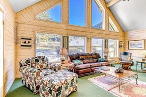 Ein gemütliches Zimmer mit Holzwänden, großen Fenstern, einem Ledersofa, einem gemusterten Hocker und einem Couchtisch aus Holz.