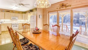 Une table à manger en bois, des chaises, un lustre, un ventilateur de plafond et une vue sur un paysage enneigé par de grandes fenêtres.