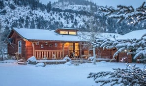 Eine Holzblockhütte mit einer Holzterrasse, umgeben von verschneiter Landschaft und Bergen.