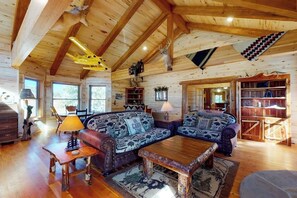 Ein rustikales Blockhaus-Interieur mit Holzbalkendecke, zwei Sofas mit gemusterten Kissen, einem Couchtisch und einem Bücherregal.