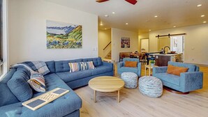 Ein Wohnzimmer mit einem blauen Sofa, einem Hocker und einem Holztisch. Es gibt einen Deckenventilator, ein Bild an der Wand und eine Küchenzeile im Hintergrund.