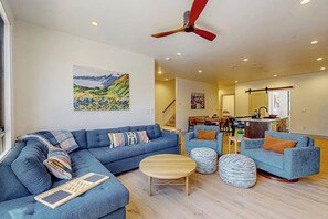 Ein Wohnzimmer mit einem blauen Sofa, einem Hocker und einem Holztisch. Es gibt einen Deckenventilator, ein Bild an der Wand und eine Küchenzeile im Hintergrund.