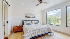 Ein Schlafzimmer mit Bett, Nachttischen, Deckenventilator, großem Fenster mit Aussicht und einem Bild an der Wand.