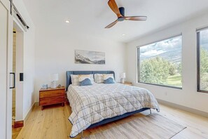 Ein Schlafzimmer mit Bett, Nachttischen, Deckenventilator, großem Fenster mit Aussicht und einem Bild an der Wand.