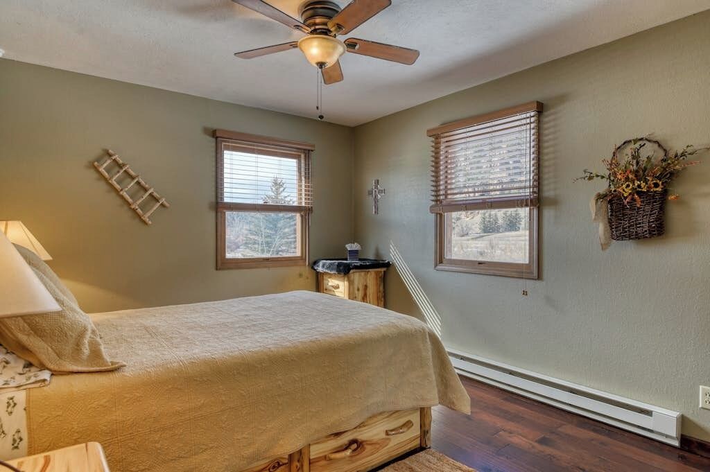 A bedroom with a bed, a ceiling fan, two windows with blinds, a wall-mounted cross, and a basket with flowers.