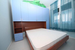 Ein Schlafzimmer mit einem Bett, einem Kleiderschrank, einer kleinen Kommode und einem Fenster mit durchsichtigen Vorhängen.