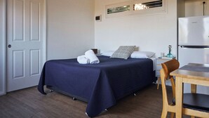 A bedroom with a bed, a wooden chair, a table, a refrigerator, and a window with blinds. - Brooman 10 - Self-Contained Cabin with Covered Deck (Mollymook)