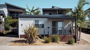 A single-story house with a covered porch, a small garden with various plants, and a clear blue sky. - Brooman 10 - Self-Contained Cabin with Covered Deck (Mollymook)