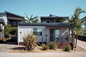 A single-story house with a covered porch, a small garden with various plants, and a clear blue sky. - Brooman 10 - Self-Contained Cabin with Covered Deck (Mollymook)
