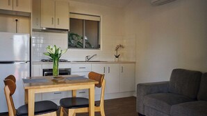 A modern kitchen with white cabinets, a dining table with chairs, a gray sofa, and a potted plant. - Brooman 10 - Self-Contained Cabin with Covered Deck (Mollymook)