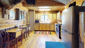 Ein Blockhaus-Interieur mit Holzwänden, einem Essbereich mit Stühlen, einer Küche mit Schränken und Geräten sowie einer Decke mit einem aufgehängten Tierkopf.