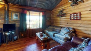 Ein Blockhaus-Interieur mit Holzboden, einer Couch, einem Fernseher und einem Fenster mit Vorhängen.