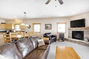 Ein Wohnzimmer mit Ledersofa, Couchtisch aus Holz, Kamin, Fernseher und Küchenzeile mit Holzschränken sowie einem Esstisch.