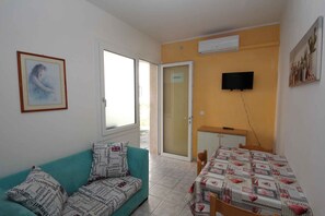 Ein Zimmer mit einer Couch, einem Esstisch, einem an der Wand montierten Fernseher und einer zur Außenseite führenden Tür.