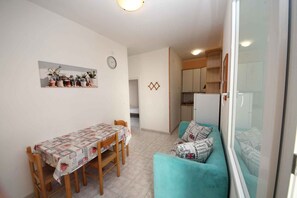 Ein Essbereich mit Tisch und Stühlen, eine Sitzecke mit gemusterten Kissen, eine Uhr an der Wand und eine Küche im Hintergrund.