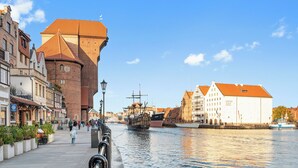 A riverside walkway with historic buildings, a tall brick tower, and a museum. - Długa Grobla 10 by Grand Apartments (Gdańsk)
