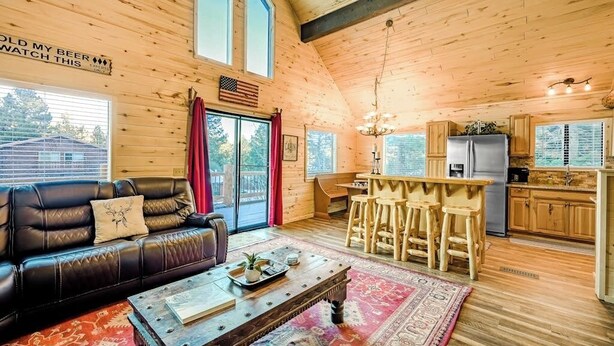 A cozy cabin interior with a leather sofa, wooden bar stools, a kitchen with a refrigerator, and a large window. - Near Town W/ Scenic Deck, Hot tub, Free WiFi, Full Kitchen & More (South Fork)