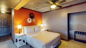 Quarto de hotel com cama, mesas de cabeceira, ventilador de teto, relógio na parede e uma porta.
