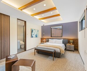 A modern bedroom with a bed, bedside tables, a bench, a mirror, and a ceiling with recessed lighting.