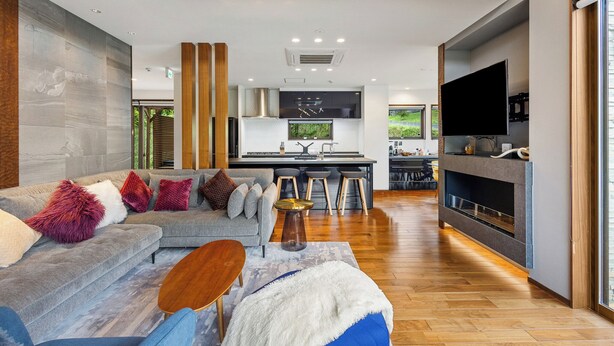 A modern living room with a grey sofa, wooden accents, and a TV. A kitchen with a large island and bar stools is visible in the background. - Mineta - Rusutsu Chalets (Rusutsu)