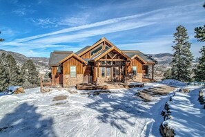 A wooden lodge with a prominent front porch, large windows, and a mountainous backdrop. - Lodge-style home with striking mountain views, fireplaces, & hot tub (South Fork)